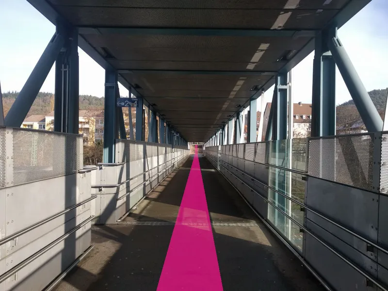 Blick durch die überdachte Fußgängerüberführung über die Gleise; ein pinker Streifen führt geradeaus weiter.